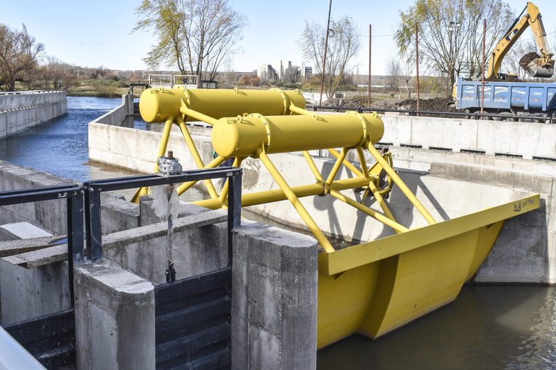 Compuerta de descarga con estructura metálica amarilla sobre un canal, con una retroexcavadora al fondo